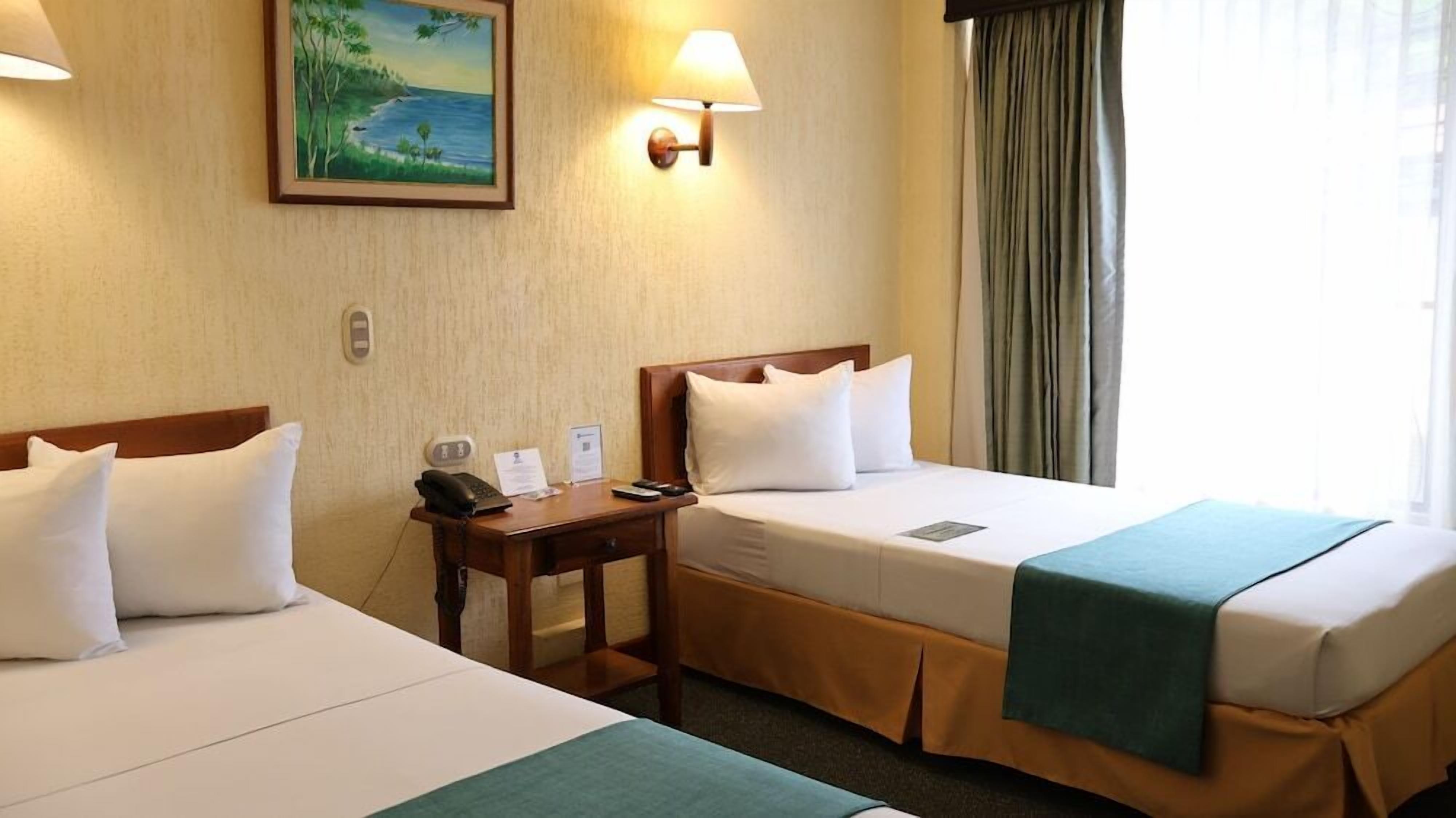 Ein Hotelzimmer mit zwei Betten, einem Schreibtisch mit Telefon und Papieren, einem Bild an der Wand und einem Fenster mit Vorhängen.