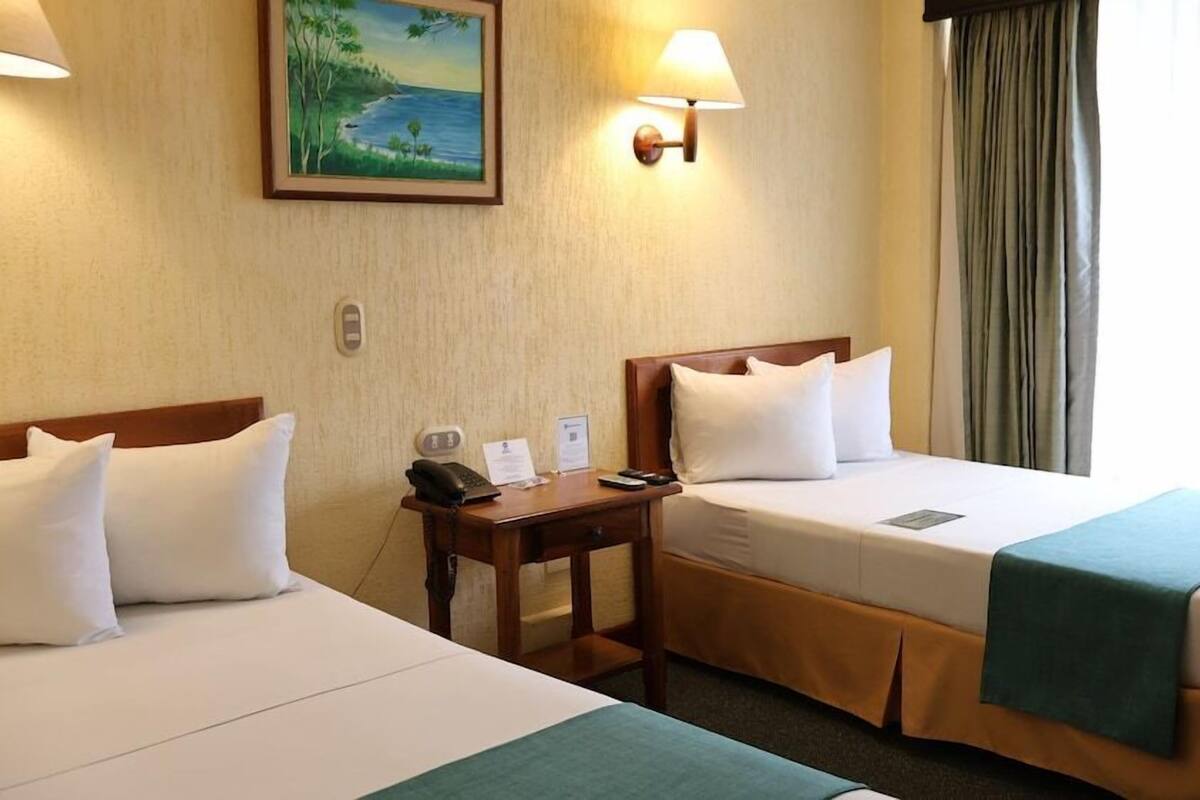 A hotel room with two beds, a desk with a phone and papers, a painting on the wall, and a window with curtains.