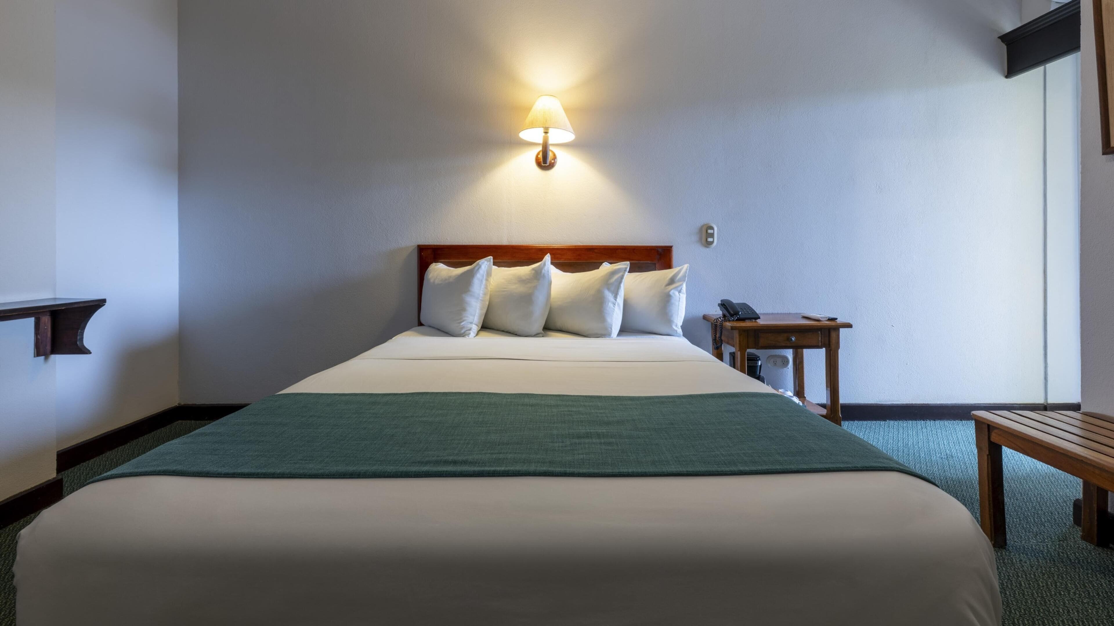A hotel room with a bed, pillows, a bedside table with a phone, and a wall-mounted lamp.