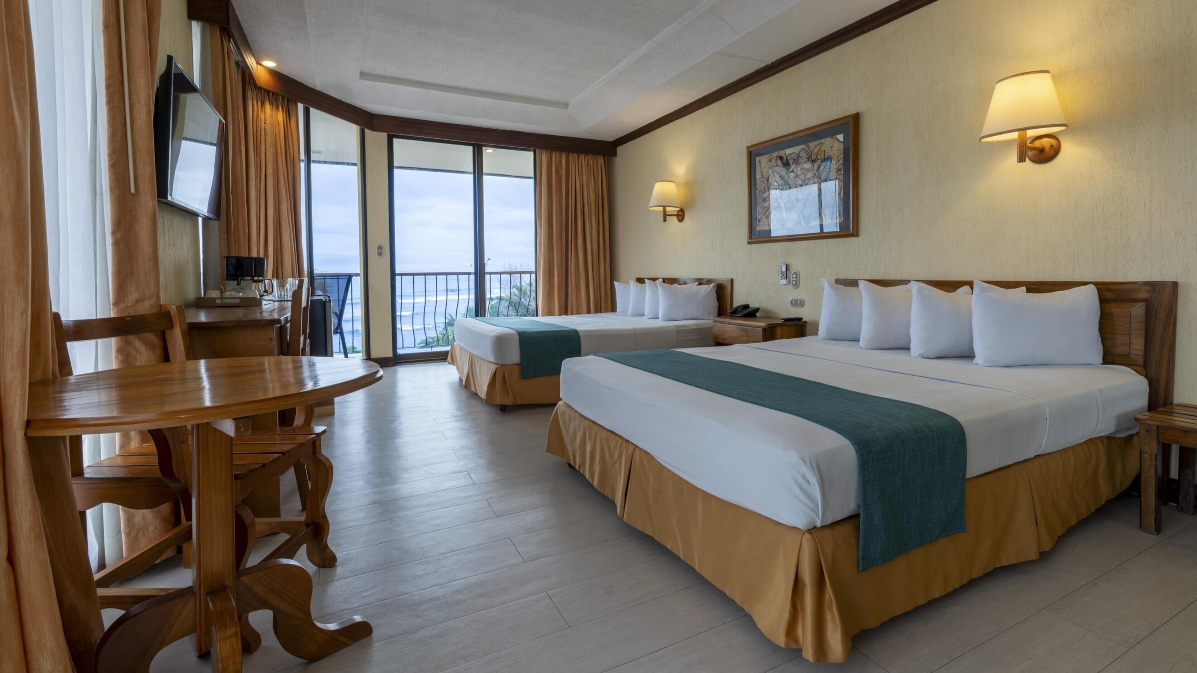 A hotel room with two beds, a dining table, and a view of the sea.