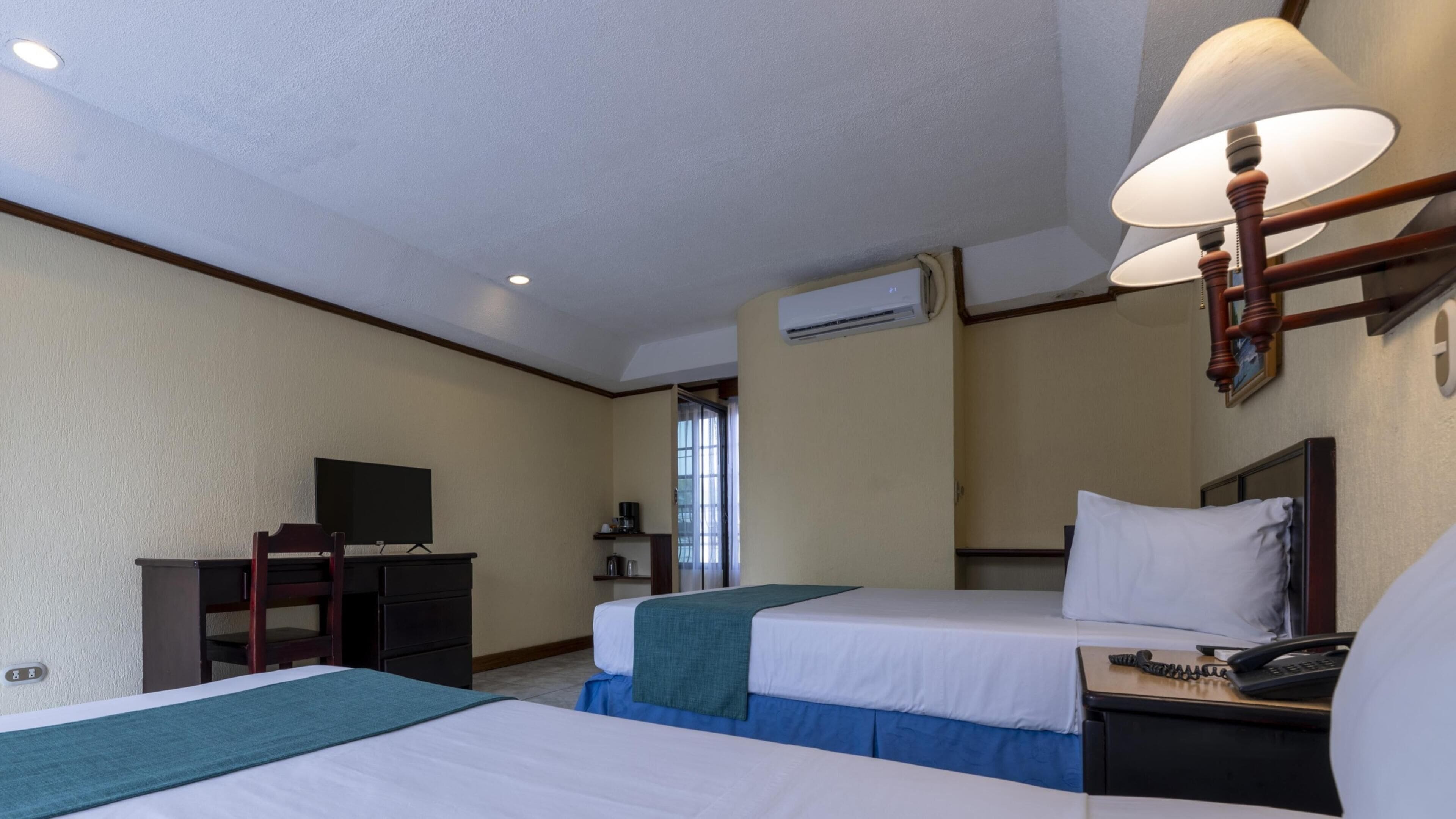 A hotel room with two beds, a desk, a television, and an air conditioning unit.