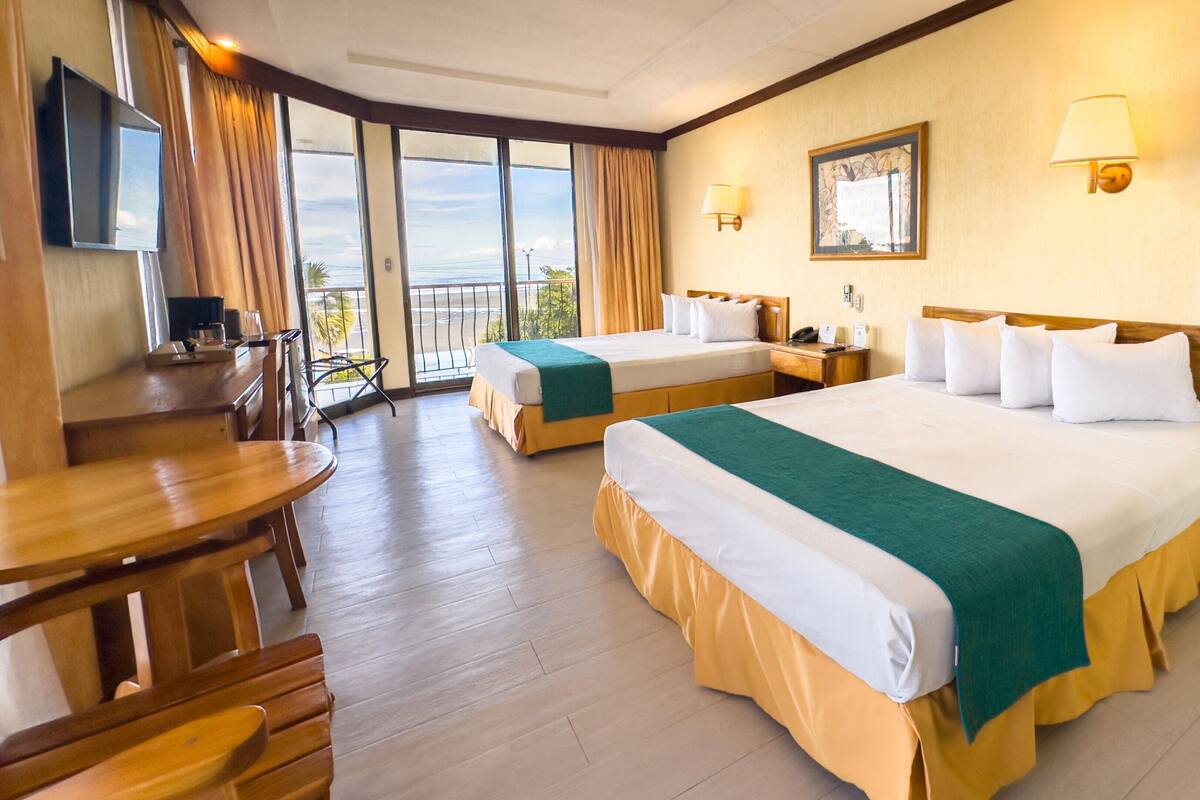 Ein Hotelzimmer mit zwei Betten, einem Schreibtisch, einem Sessel, einem Fernseher und einem Balkon mit Blick.