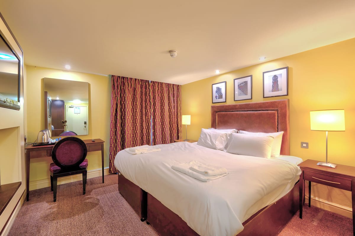 A hotel room with a large bed, a desk with a chair, a mirror, and a lamp.