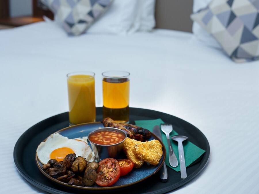 A breakfast tray with eggs, beans, tomatoes, and toast on a plate, accompanied by two glasses of orange juice, set on a bed with a headboard and pillows in the background.