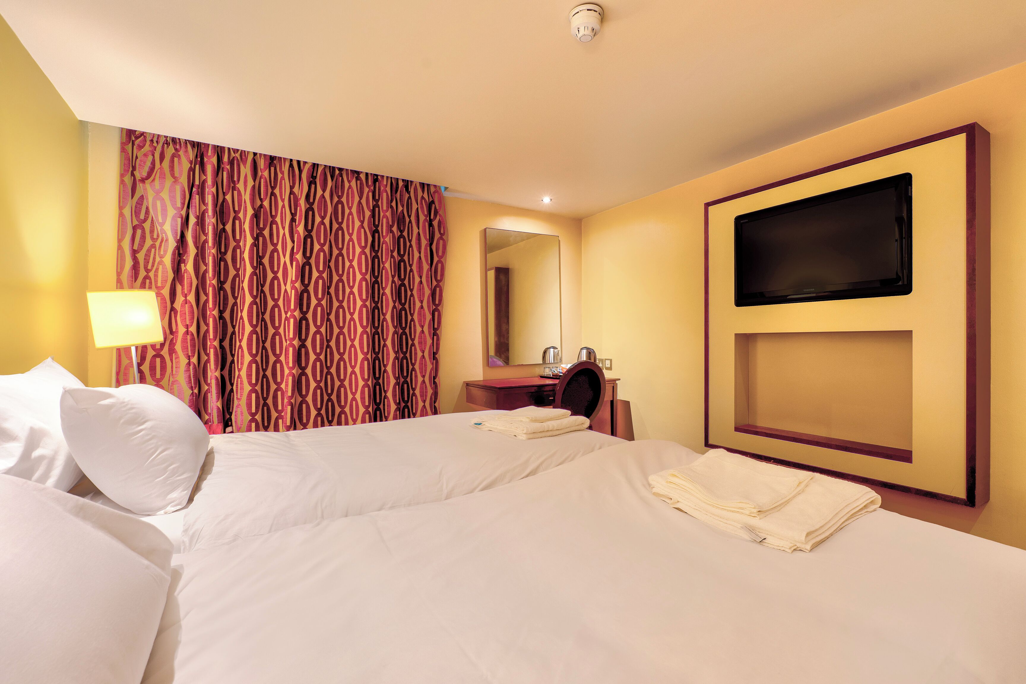 Habitación de hotel con dos camas, televisión, espejo, lavabo y cortina estampada.