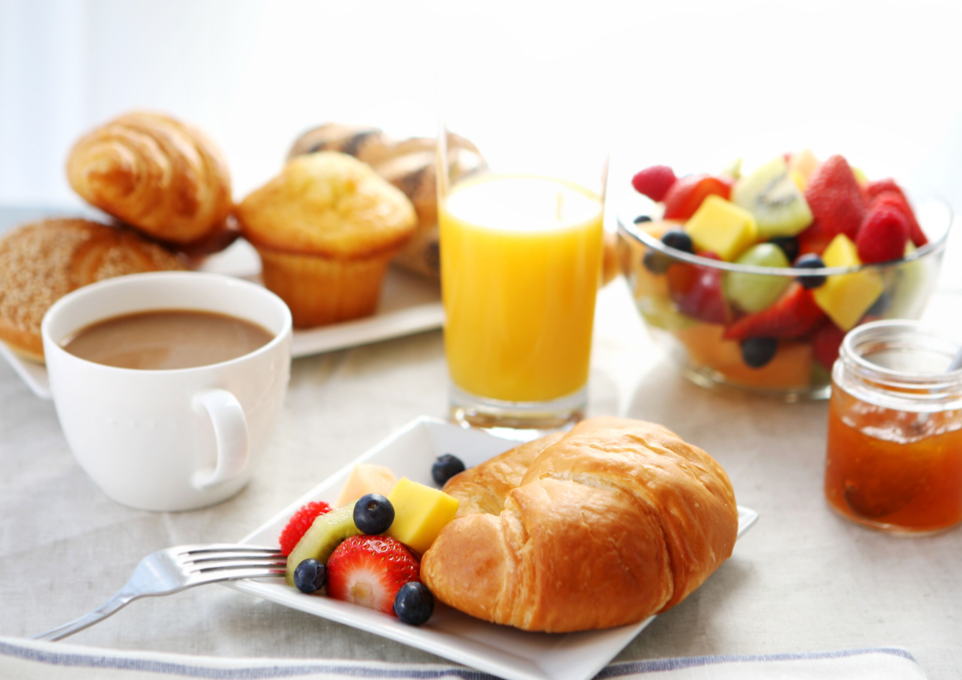 A breakfast setting with croissants, a fruit bowl, a glass of orange juice, a cup of coffee, and a jar of jam.