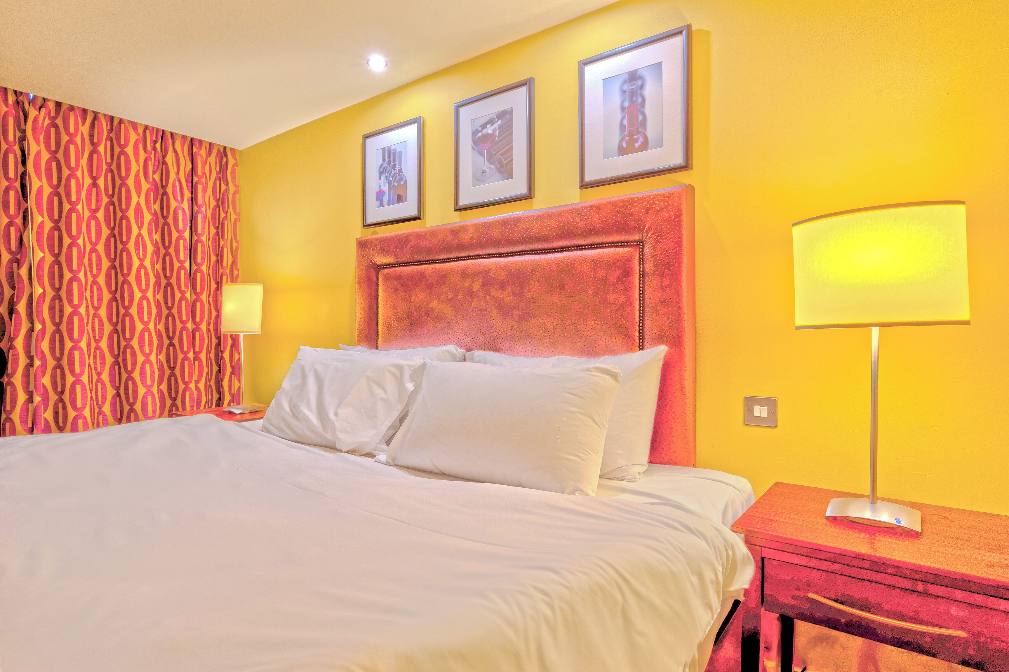 Una habitación de hotel con una cama grande, una mesita de noche de madera, una lámpara y tres cuadros en la pared.