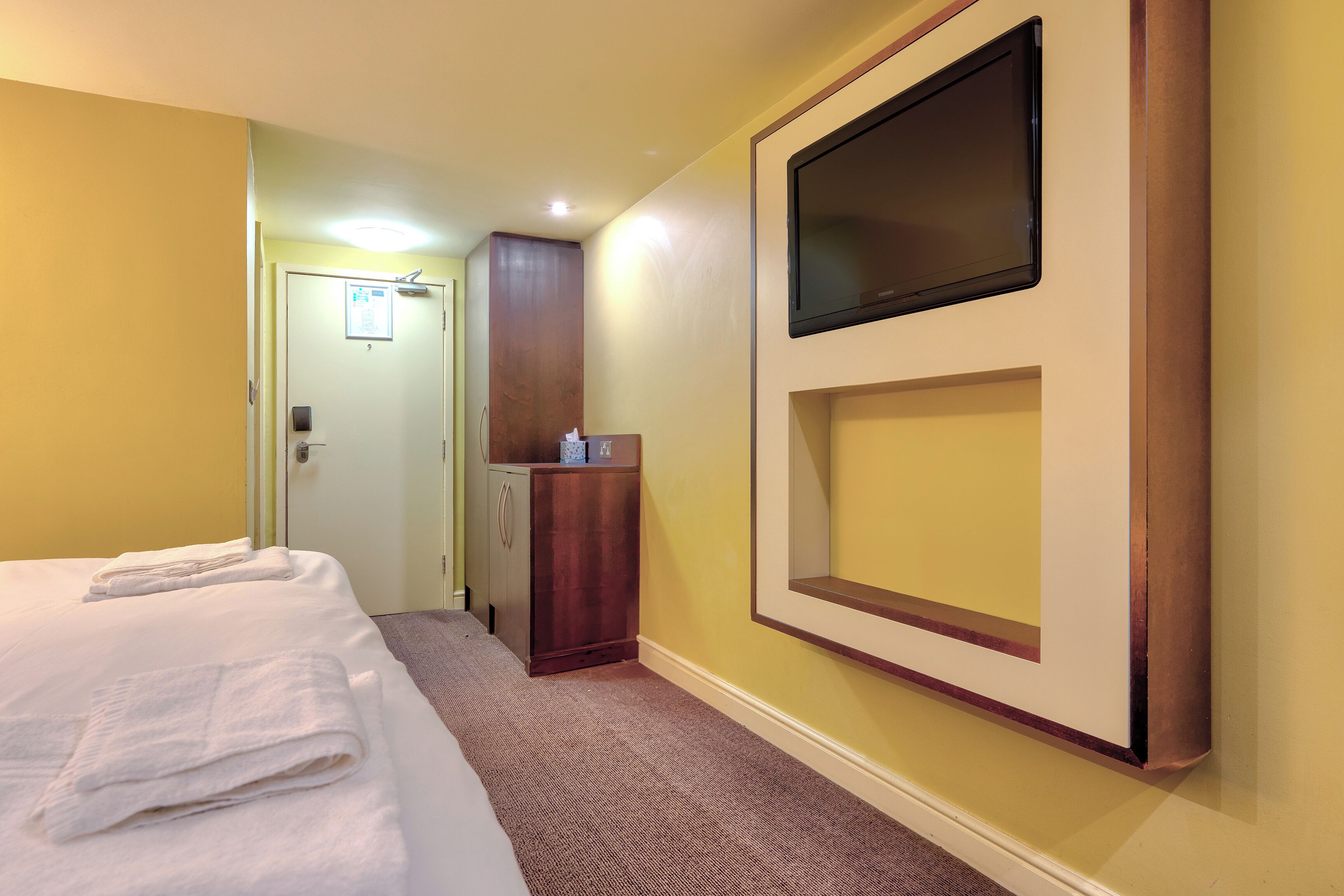 A hotel room with a bed, a wooden cabinet, a flat-screen TV mounted on the wall, and a door with a sign.