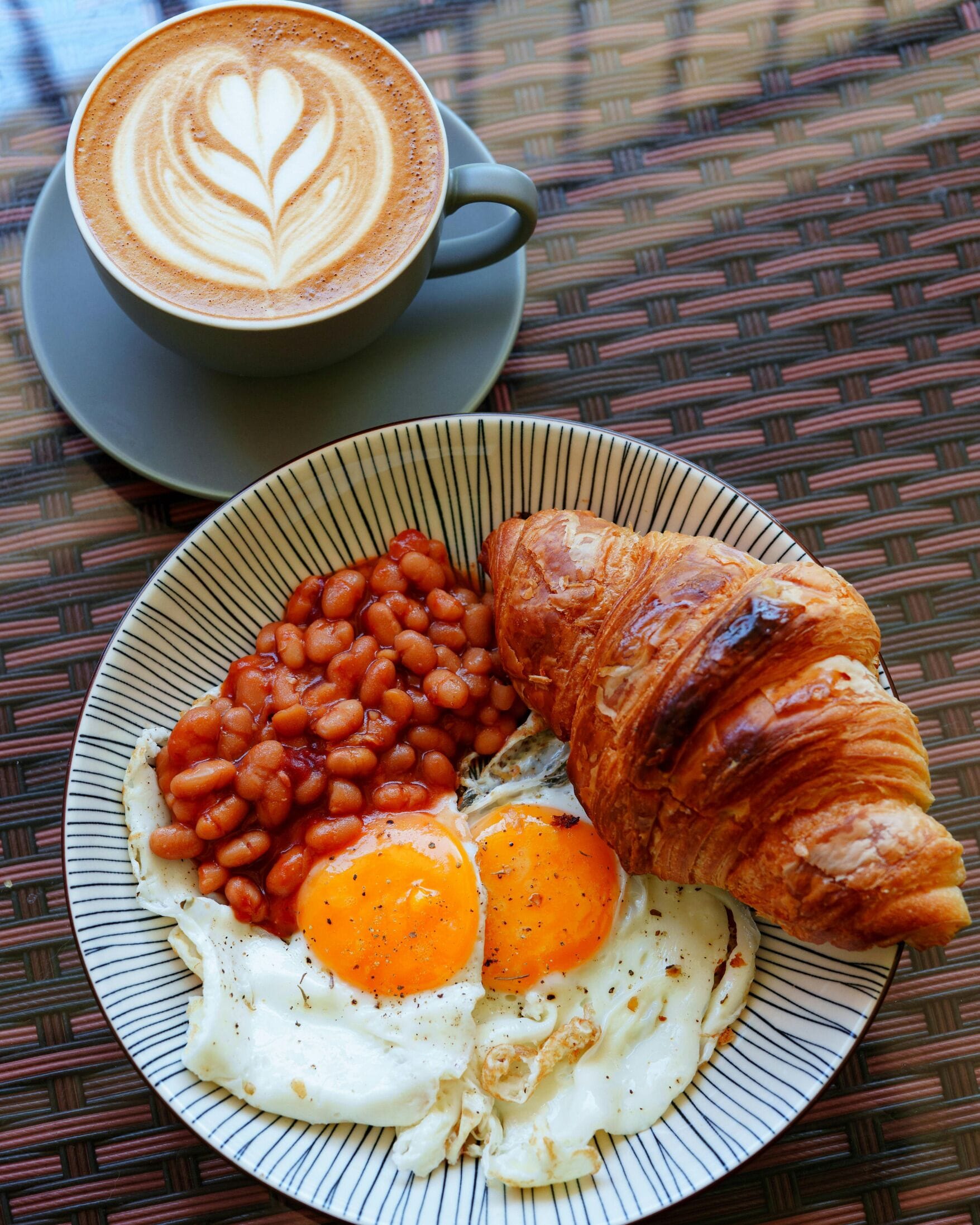 Un plato con huevos, frijoles y un croissant, junto a una taza de café sobre un mantel tejido.