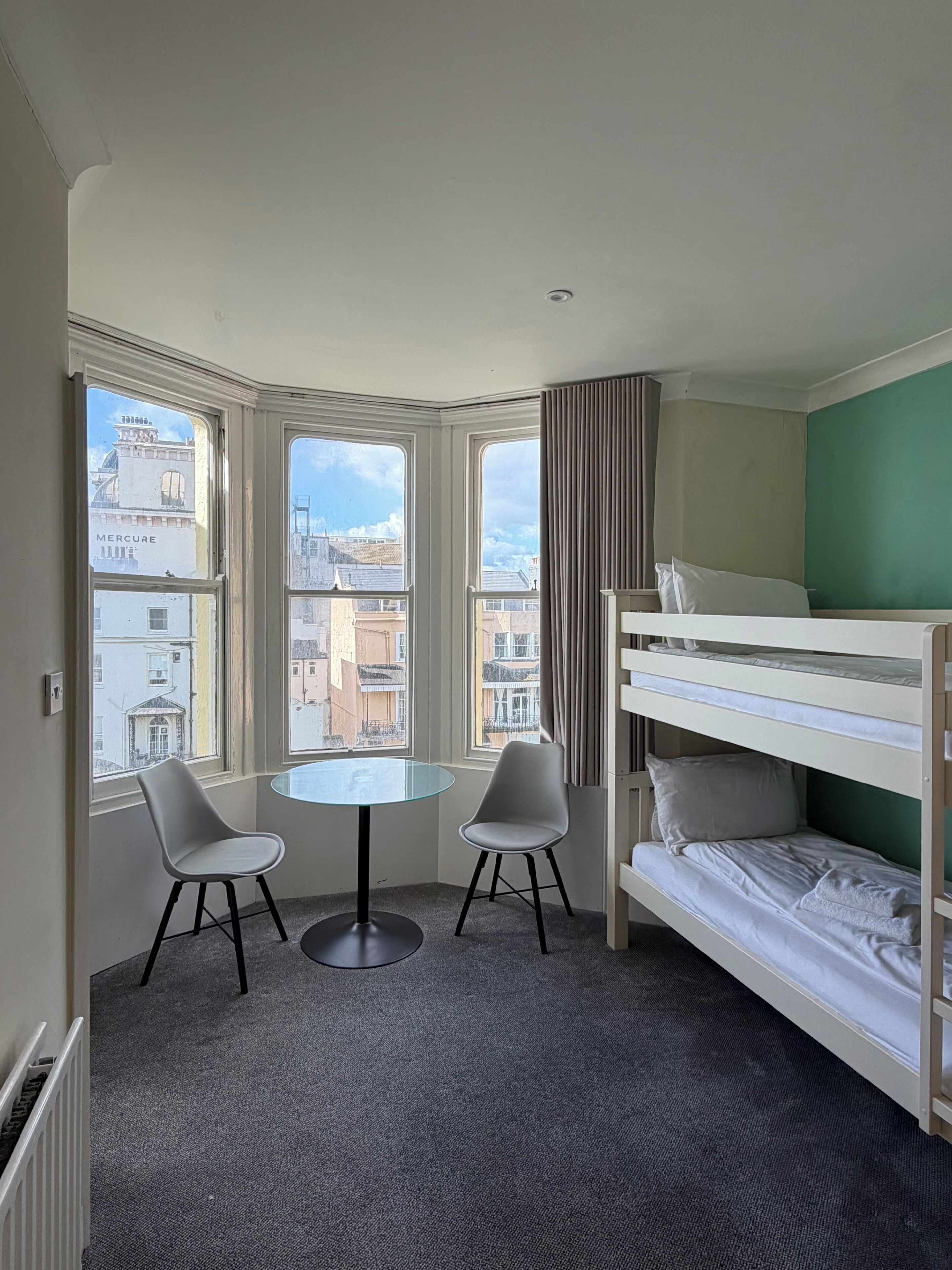 Una habitación con literas, dos sillas, una mesita y ventanas amplias con vistas a la ciudad.