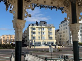 Um edifício com vários andares, janelas salientes, passadeira para pedestres e rua com carros e pessoas.