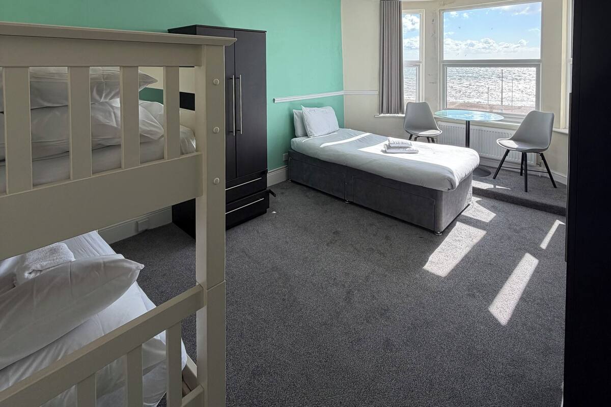A hotel room with a bunk bed, a single bed, a desk with chairs, a window with a view, and a radiator.