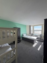 A hotel room with a bunk bed, a single bed, a desk with chairs, a window with a view, and a radiator.