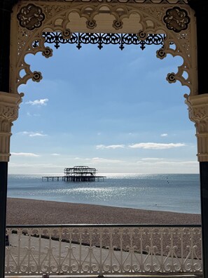 Vista de la playa con un muelle que se adentra en el mar, cielo despejado y una barandilla metálica decorativa.