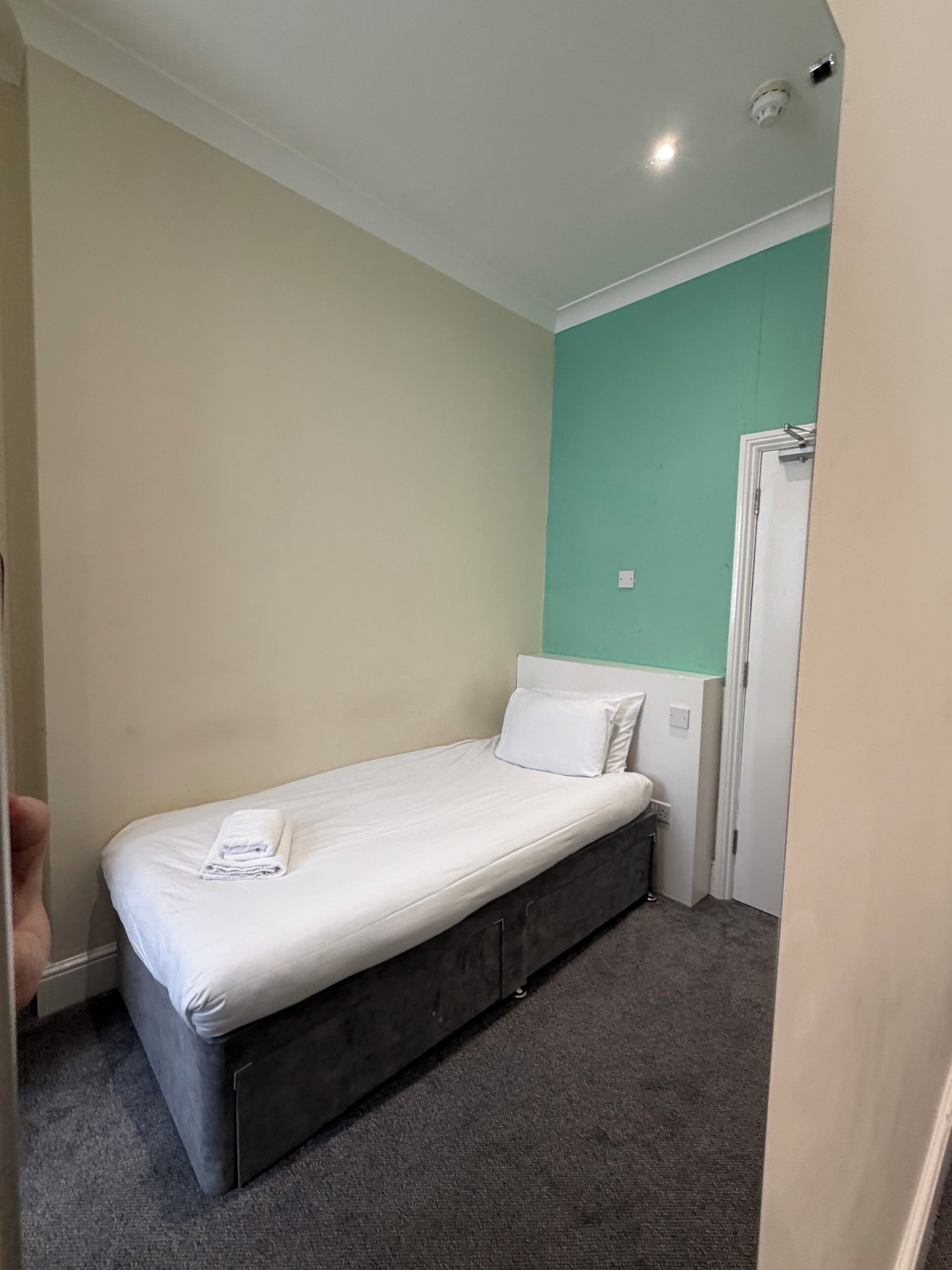 A small, single-bed room with a grey carpet, a white mattress, and a teal accent wall.