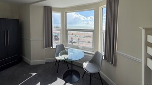 A room with a bay window, a dining table with chairs, a black cabinet, and a view of the beach and sea.