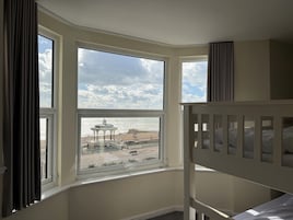 A room with a view of the beach, a white bunk bed, and a large window with curtains.