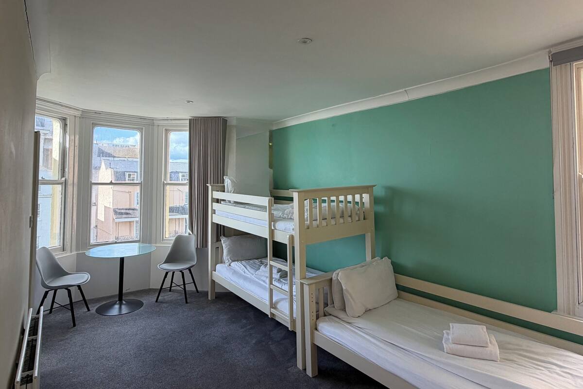 A room with bunk beds, a small table, two chairs, and a view of buildings through the window.