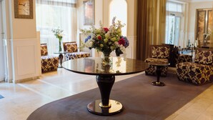 Mesa redonda com um vaso de flores no centro, rodeada por sofás estampados e uma janela grande com cortinas.