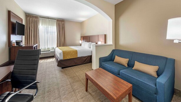 Habitación de hotel con una cama, un sofá azul, una mesa de centro de madera, un escritorio con una silla, un televisor montado en la pared y una ventana con cortinas.