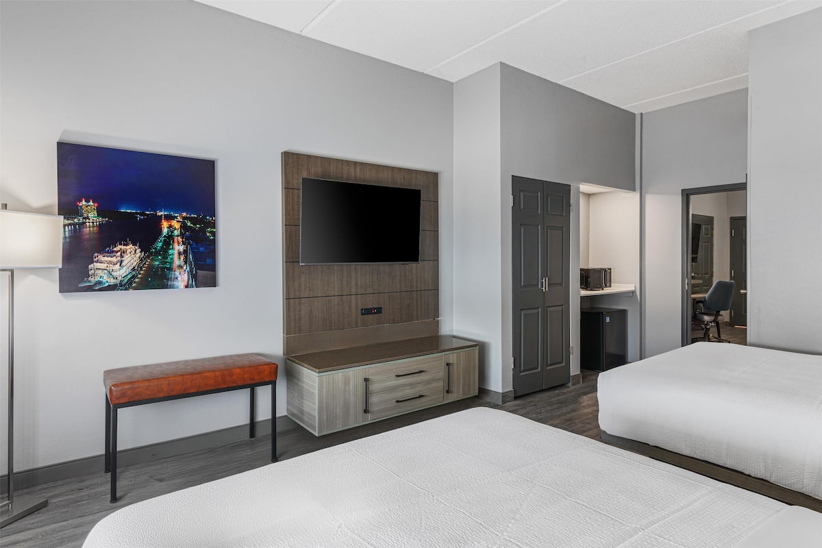 A hotel room with a large flat-screen TV mounted on a wooden cabinet, a bench, two beds, a desk, and a wardrobe.