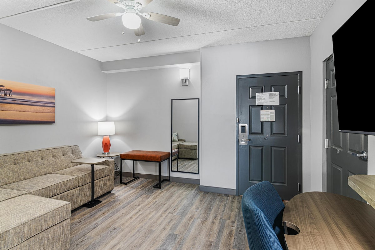 A hotel room with a sofa, a small table with a lamp, a wooden desk, a chair, a ceiling fan, and a door with a sign.