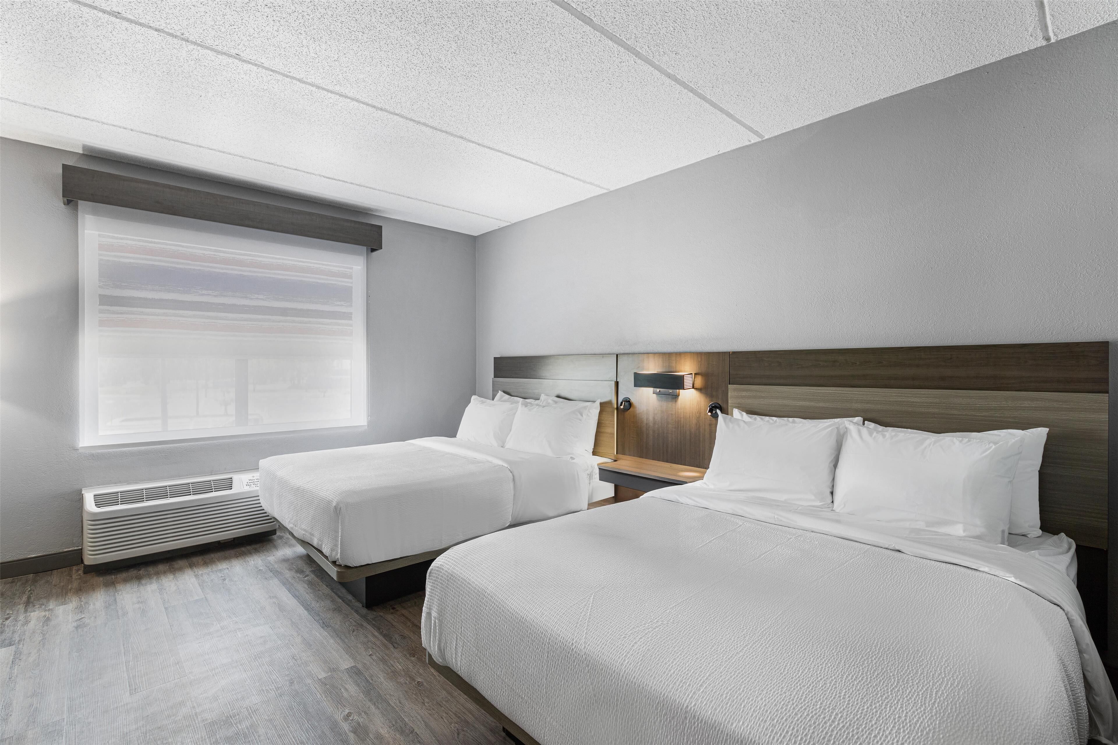 Habitación de hotel con dos camas, una ventana con persianas, un calentador en la pared y una mesita de noche con lámpara.