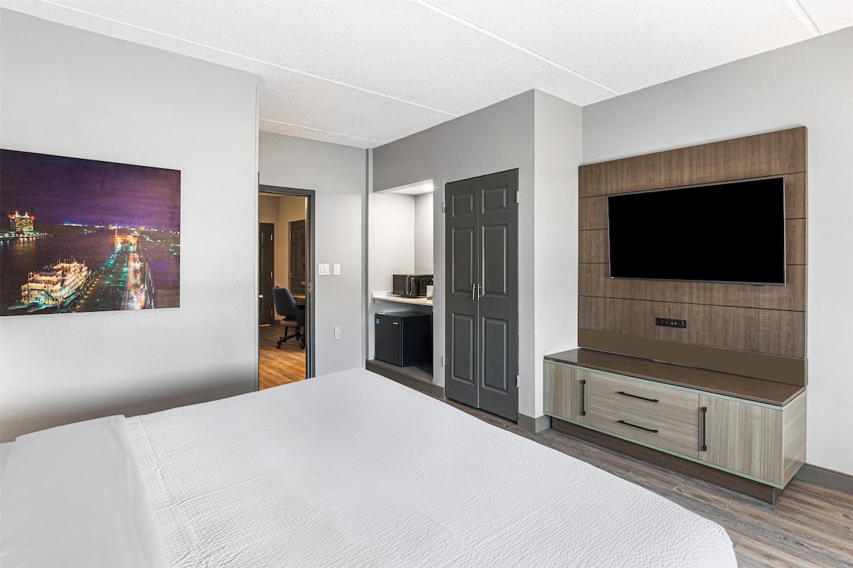 A modern hotel room with a large bed, a flat-screen TV mounted on a wooden panel, a small kitchenette, and a door leading to another room.