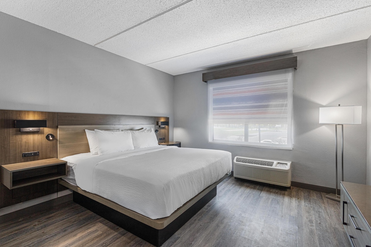 A modern hotel room with a large bed, built-in nightstands, a window with blinds, a floor heater, and a bedside lamp.