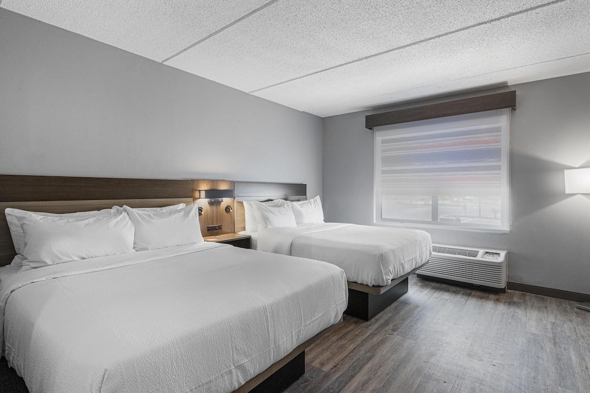 A hotel room with two beds, a nightstand, a window with blinds, and a ceiling vent.