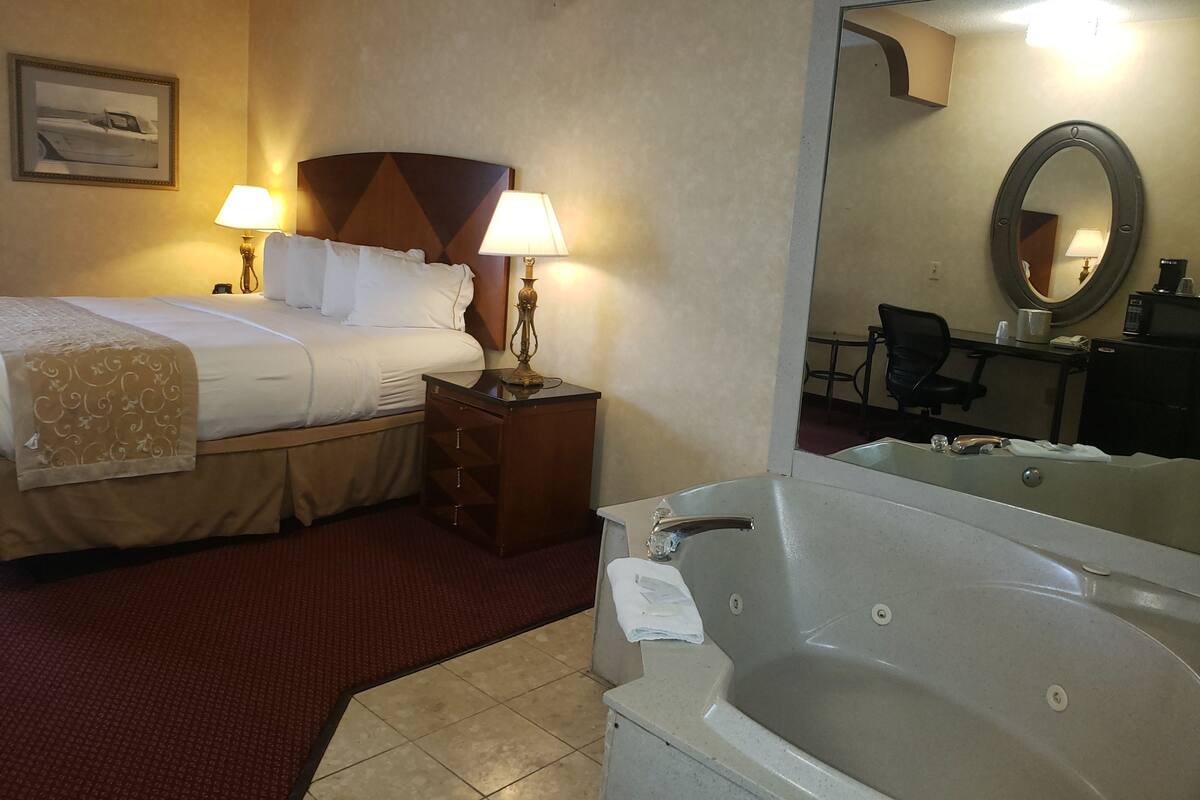 Quarto de hotel com cama, duas luminárias de cabeceira, mesa de cabeceira, escrivaninha com cadeira, espelho grande e banheira.