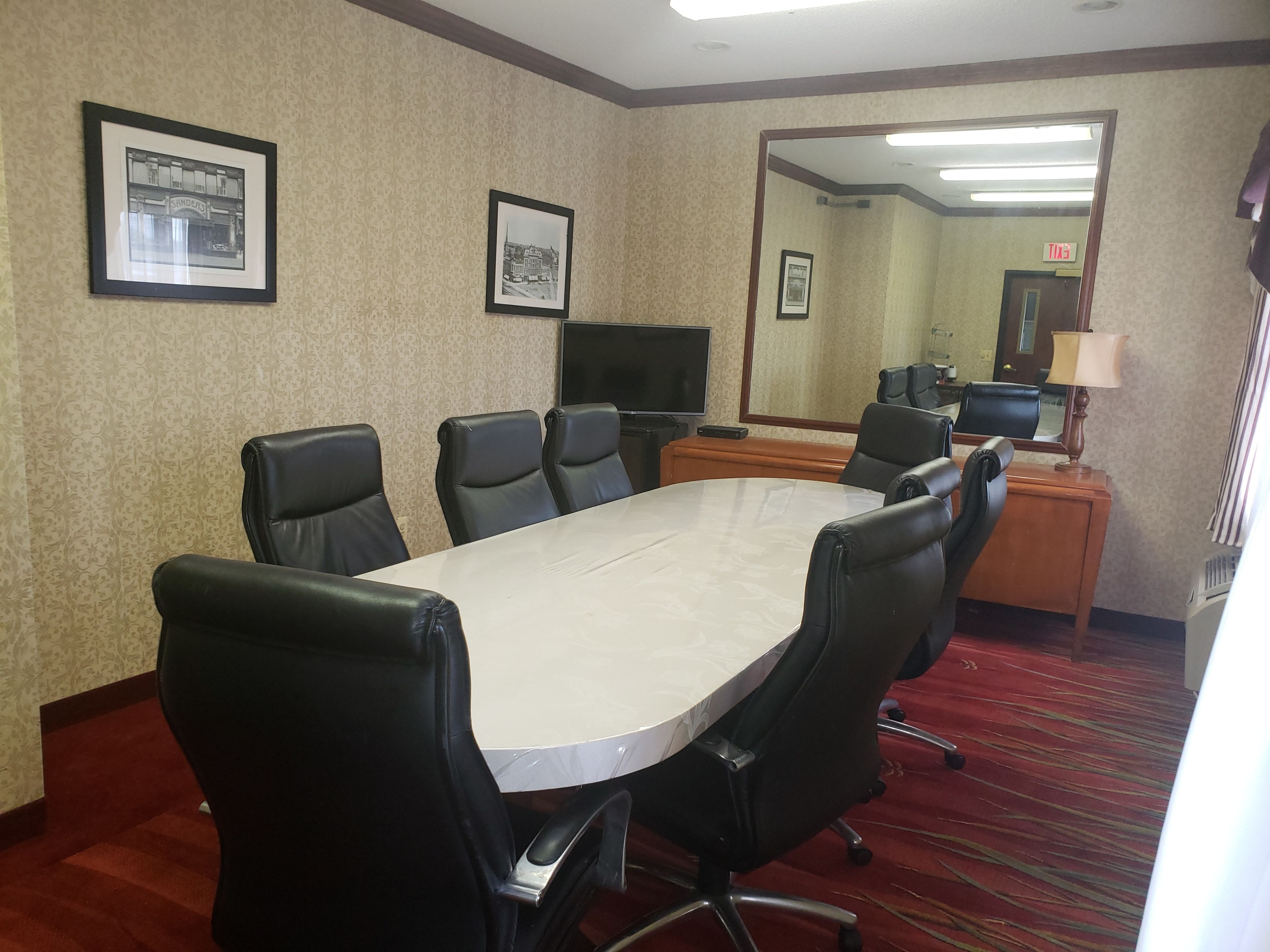 A conference room with a large table, black leather chairs, framed pictures on the walls, a desk with a computer, and a mirror reflecting the room.
