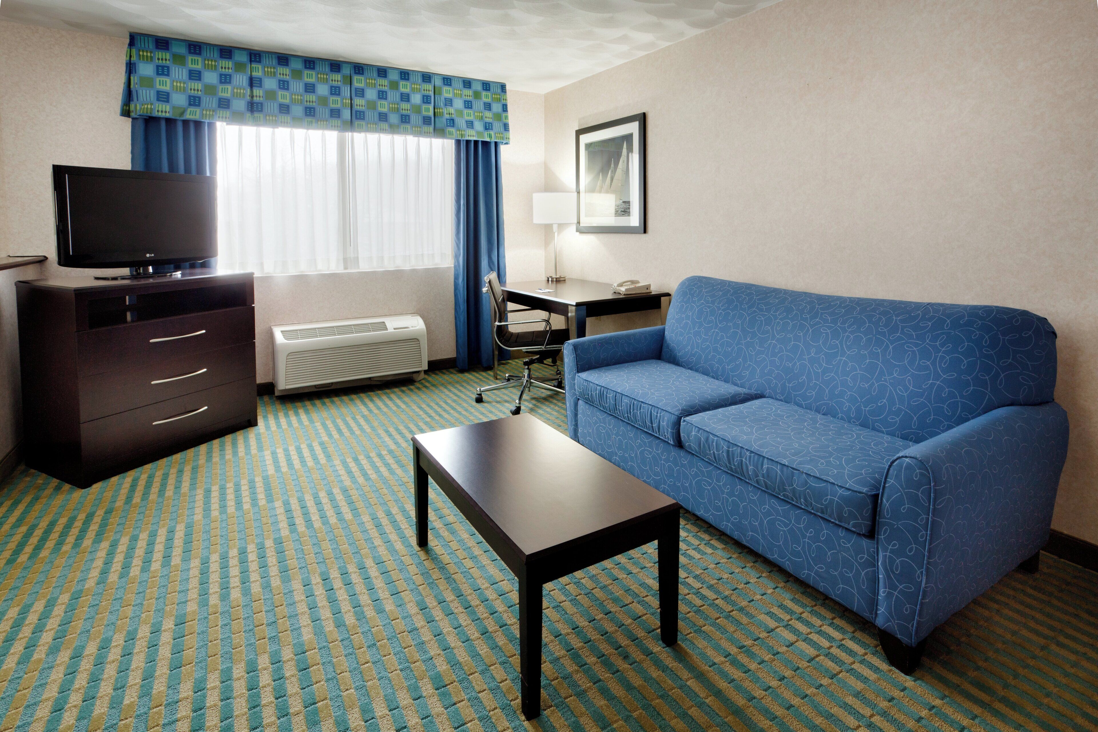 A hotel room with a blue sofa, a TV, a desk, and a chair.