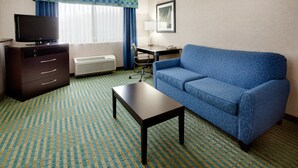 A hotel room with a blue sofa, a small table, a TV on a dresser, a desk, and a chair. - Holiday Inn Express & Suites Smithfield - Providence by IHG (Smithfield)