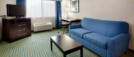 Habitación de hotel con un sofá azul, una mesita, un televisor sobre un aparador, un escritorio y una silla.