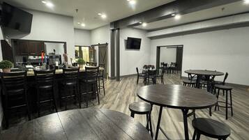 A modern kitchen with dark wooden cabinets, a built-in oven, and a microwave. There is a dining area with round tables and chairs, and a large wooden table in the center.