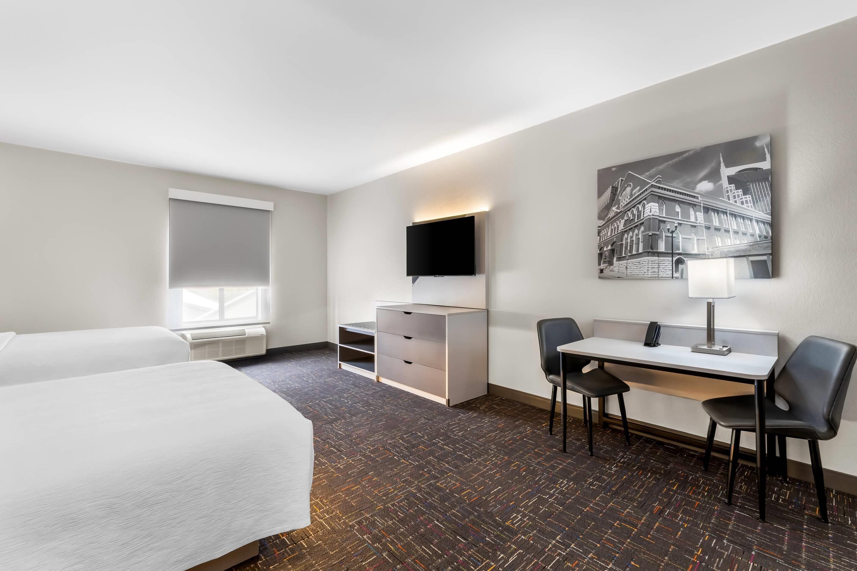 Une chambre d’hôtel avec deux lits, un bureau avec une chaise, un téléviseur à écran plat, une lampe de bureau et un tableau au mur.
