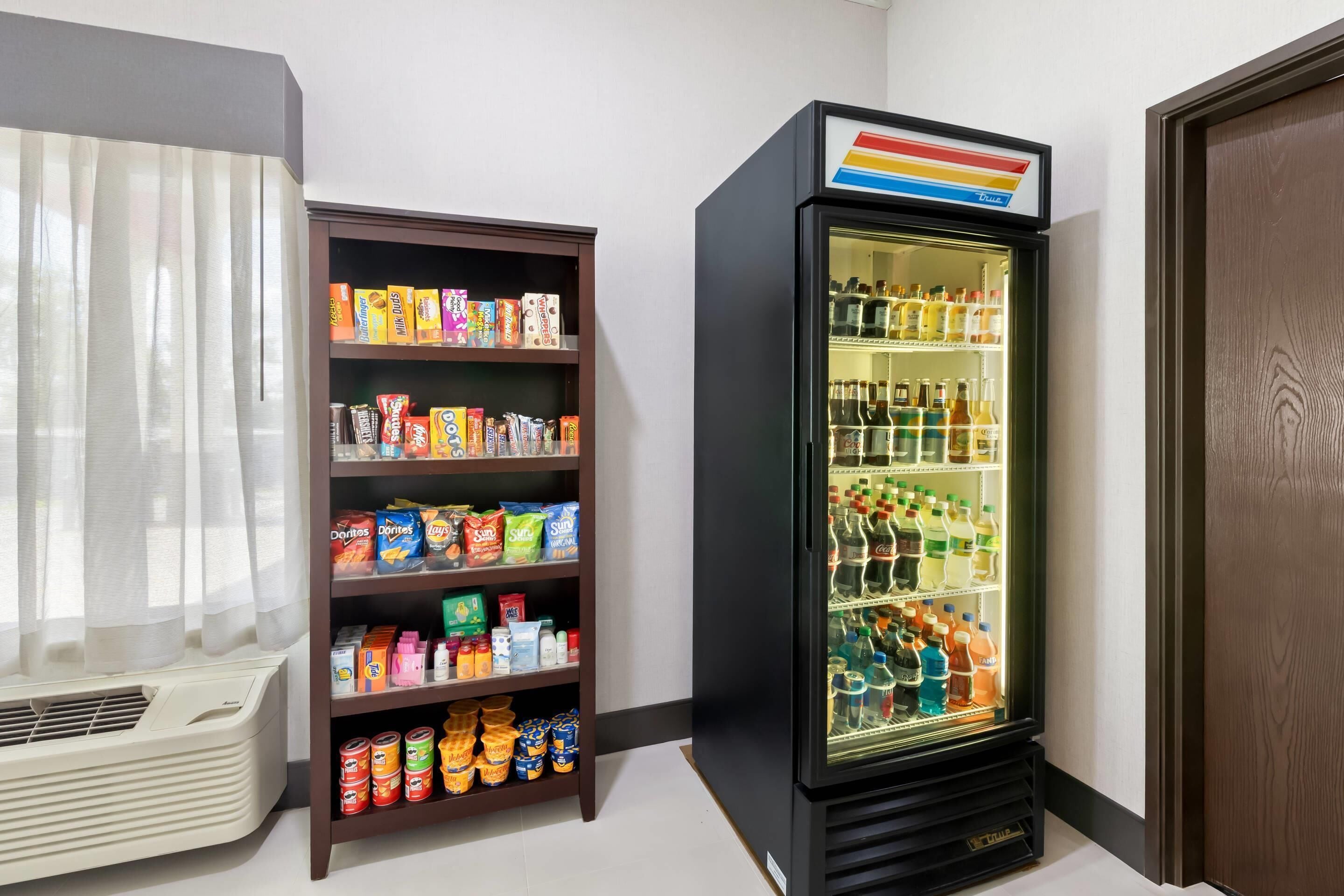 A room with a snack shelf stocked with various packaged foods and a beverage refrigerator containing multiple bottles and cans.