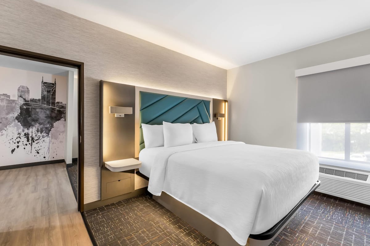 A modern hotel room with a large bed, a nightstand, a cityscape artwork on the wall, and a window with a view.