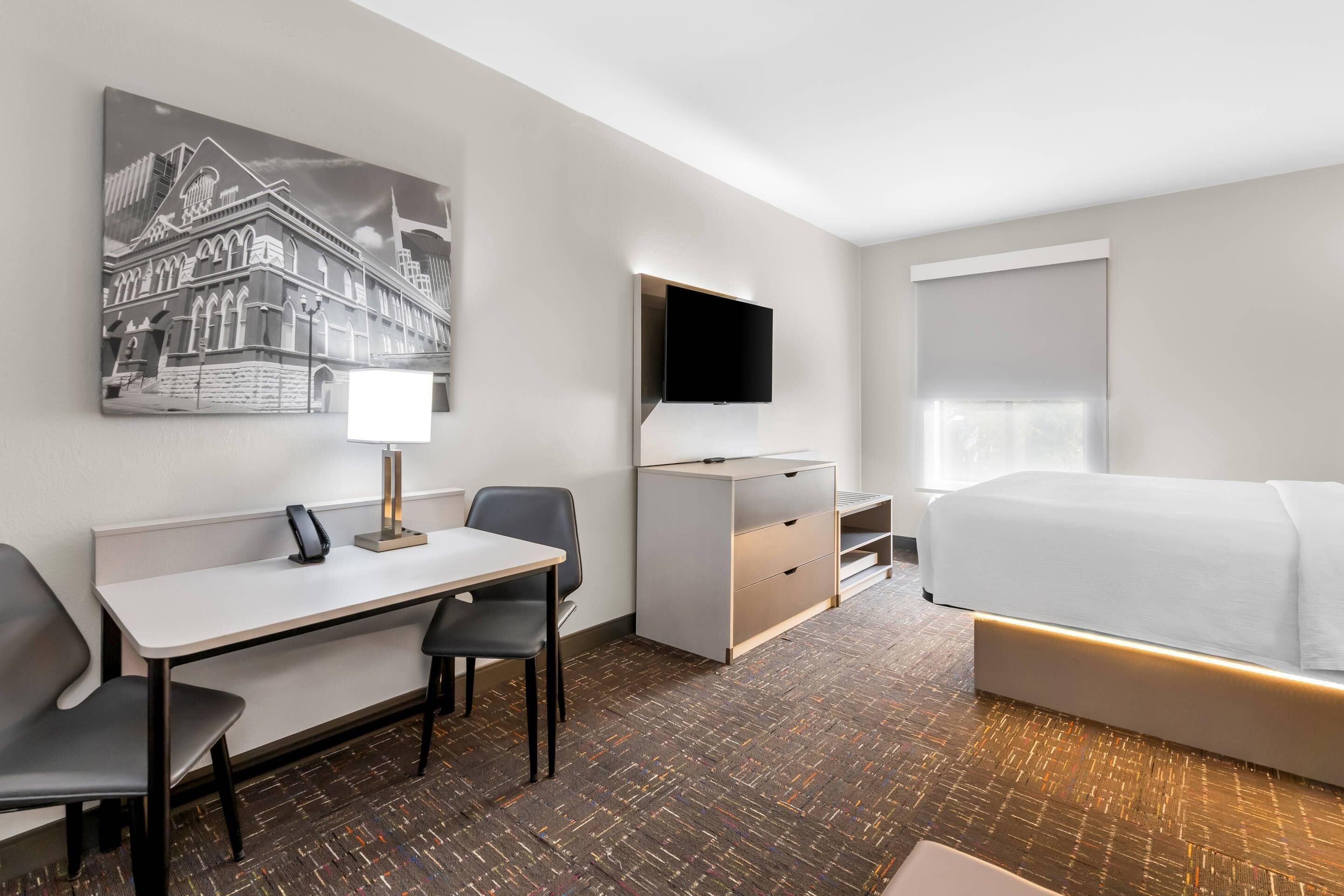 A hotel room with a bed, a desk with a chair, a flat-screen TV, a bedside table, and a large window with blinds.