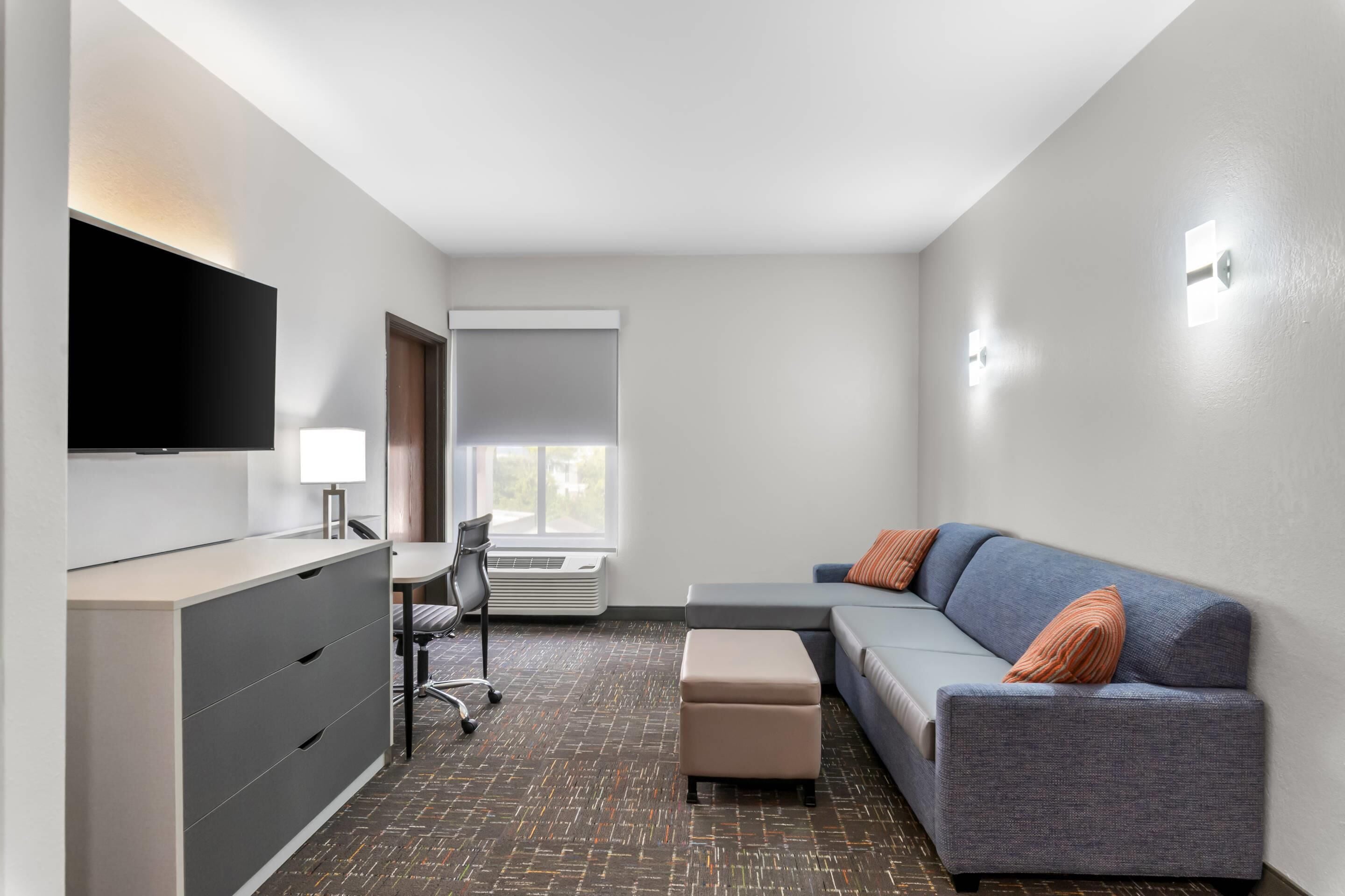 Une chambre d’hôtel moderne équipée d’une télévision à écran plat, d’un bureau avec une chaise, d’un canapé avec des coussins et d’une grande fenêtre laissant entrer la lumière naturelle.