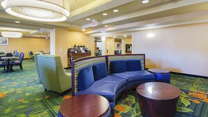 A hotel lobby with a curved blue sofa, a green patterned armchair, and a wooden coffee table. There are multiple tables and chairs, and an exit sign is visible on the wall. - Quality Inn Boston - Revere (Revere)
