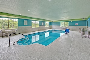 An indoor swimming pool with clear pool rules posted, a lifeguard chair, and a view of the outdoors through large windows.