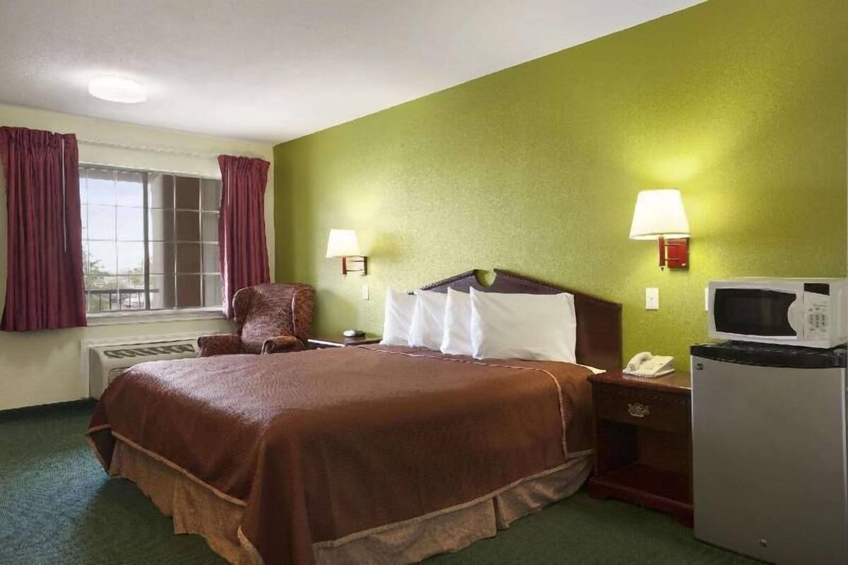 A hotel room with a bed, a chair, a desk with a lamp, a microwave, and a telephone.