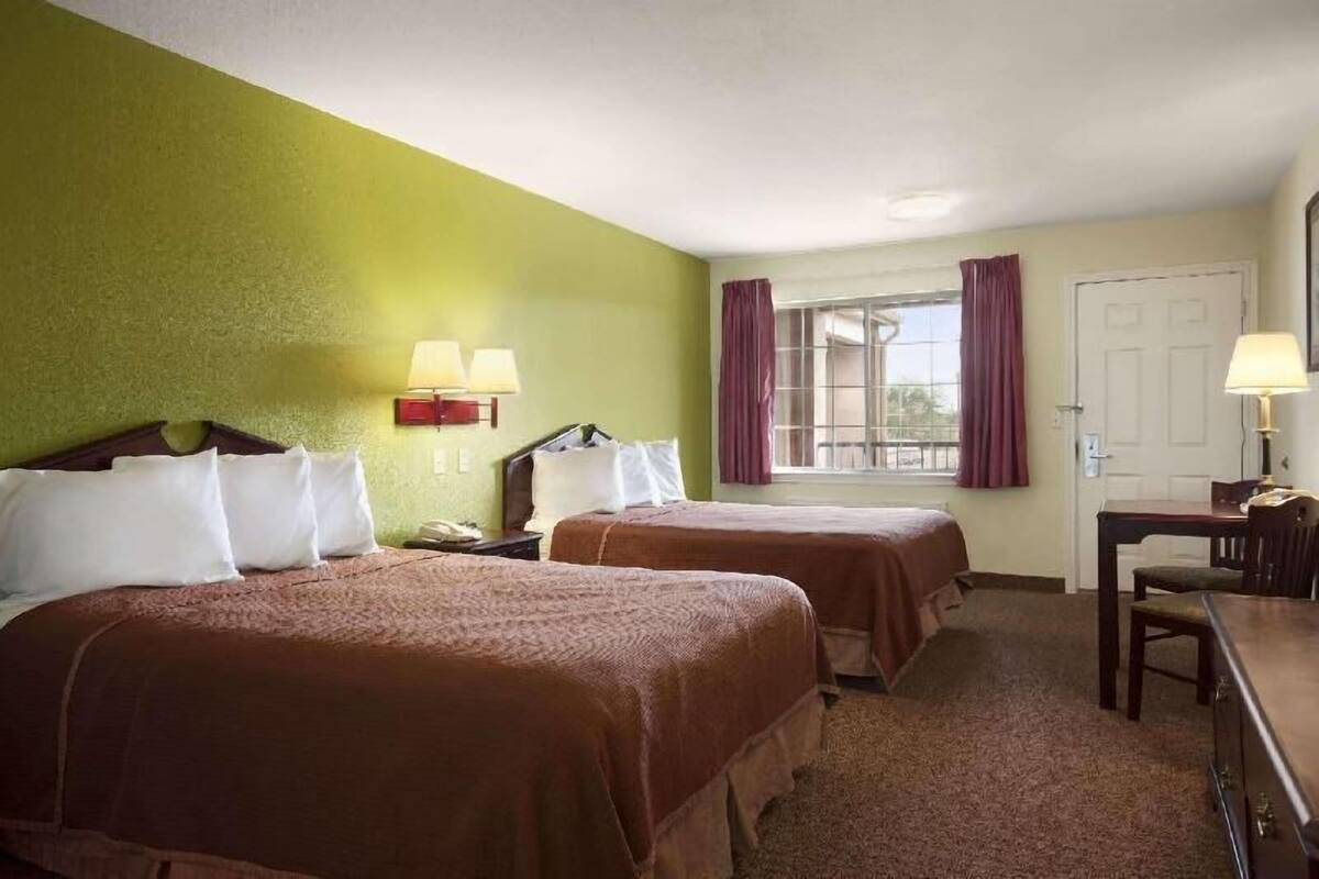 A hotel room with two beds, a desk, a chair, a window with curtains, and a door.