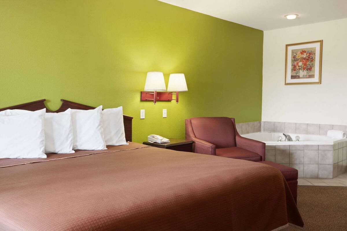 A hotel room with a large bed, a chair, a desk with a telephone, a wall-mounted lamp, a painting, and a bathtub.