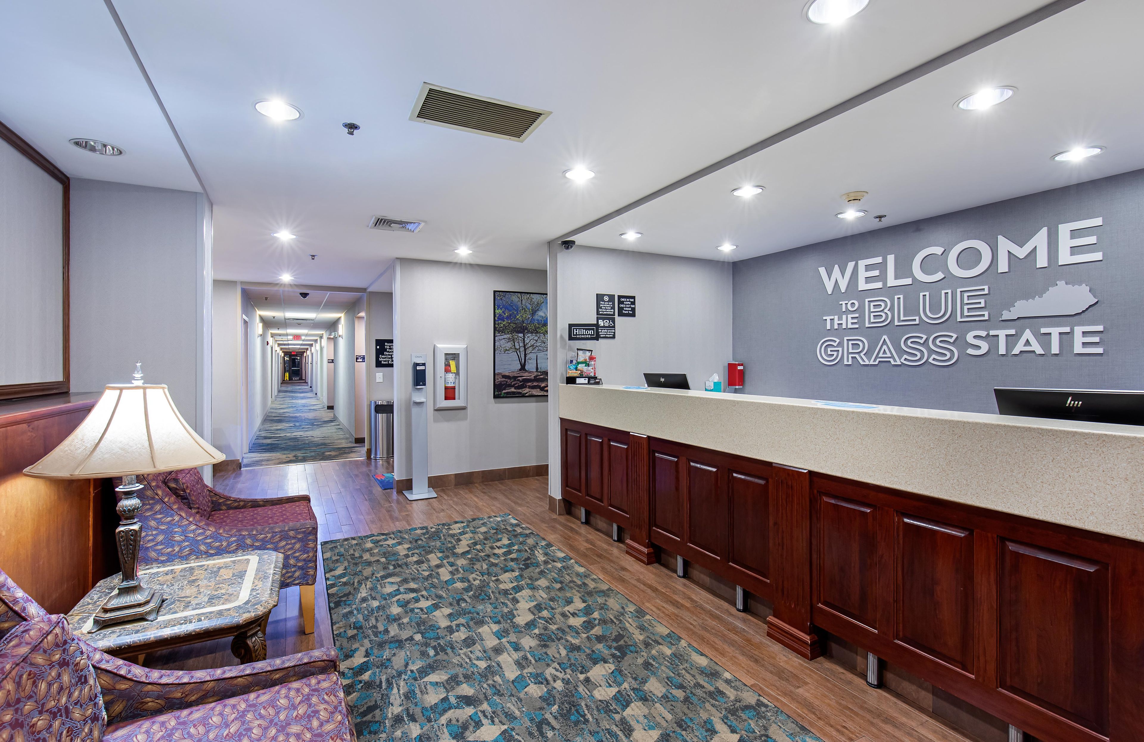 A hotel lobby with a reception desk, a carpeted area with a sofa, and a hallway leading to the elevators.