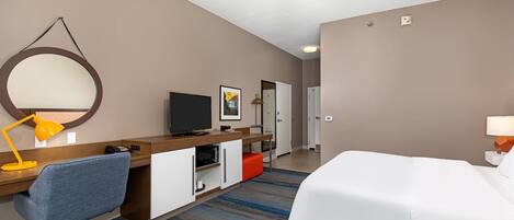 Habitación de hotel con cama, escritorio, silla, televisor y un baño visible al fondo.