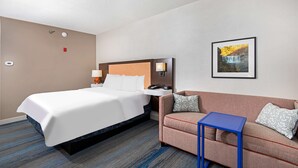 Une chambre d’hôtel comprenant un lit, un canapé, une petite table, une lampe fixée au mur, un tableau encadré et un thermostat.