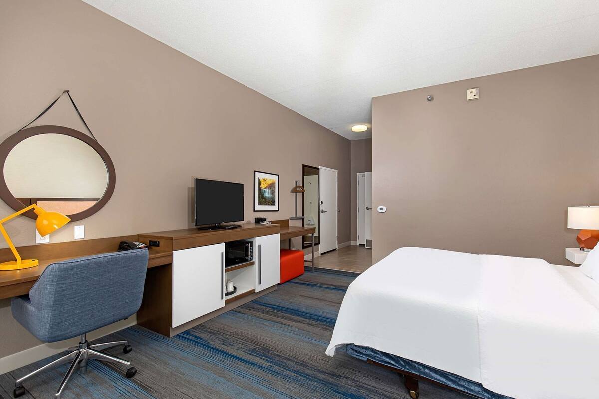 A hotel room with a bed, desk, chair, television, and a large mirror.